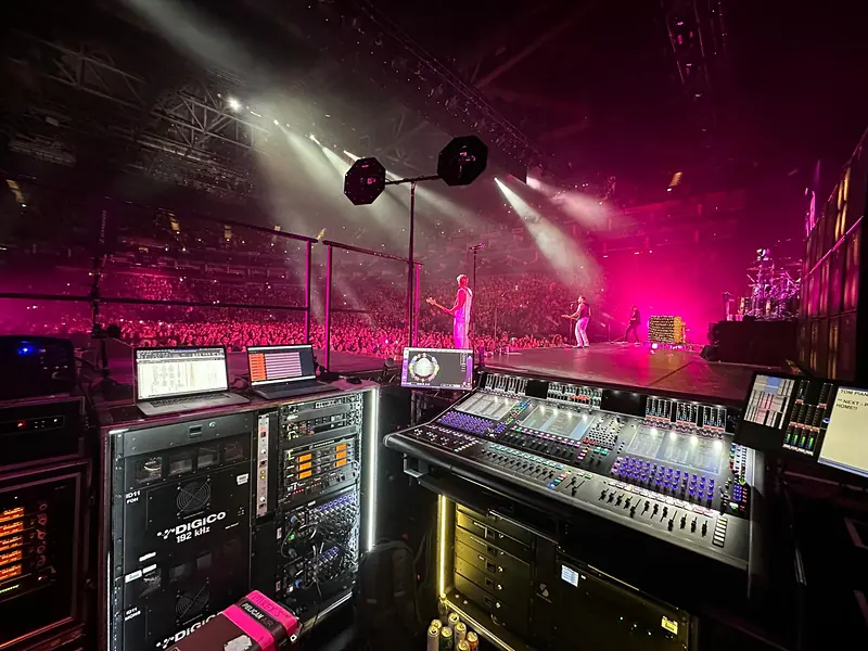 DiGiCo console, racks and laptops beside a pink-lit arena stage with the band performing