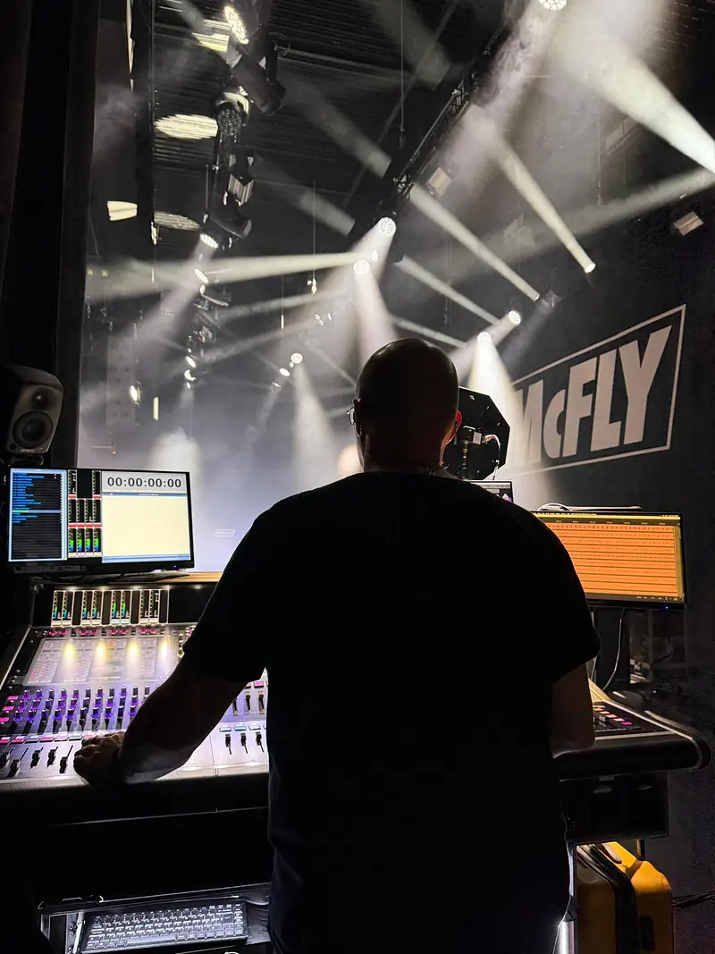 Mike Osman mixing at a DiGiCo console, seen from behind, with beams of light and a McFly sign beyond